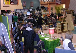 People attending an indoor event with multiple informational booths, including displays, seating areas, and tables, in a busy, decorated space.