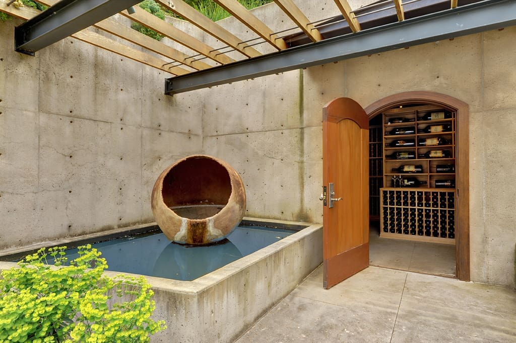 An open door reveals a wine cellar with numerous wine bottles on wooden shelves. Outside, there is a water feature with a large, hollow, spherical sculpture in a shallow pool.