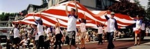 A group of people in matching shirts holds up a large American flag, parading along a street lined with spectators.