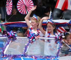 Two children wearing 4th of July-themed accessories wave from a decorated float in a parade, surrounded by patriotic decorations and an inflatable figure in the background.