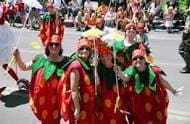 A group of people dressed in strawberry costumes stands together in a parade, with a crowd of spectators in the background.