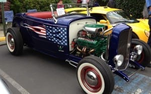 A blue vintage hot rod with an American flag design on the side, equipped with a green engine and whitewall tires. A yellow car is parked nearby.