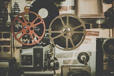 A vintage film projector with two large reels is displayed against a wall with other nostalgic items, including a vinyl record and an old photograph.