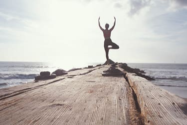 Silhouetted person practices yoga in tree pose on a wooden pier extending into the ocean with a cloudy sky in the background.