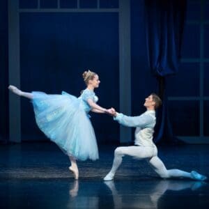 Two ballet dancers perform on stage; the female dancer balances on pointe in a light blue tutu while the male dancer kneels, holding her hands.