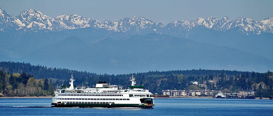 A ferry boat sails on a calm body of water with forested land and snow-capped mountains in the background.