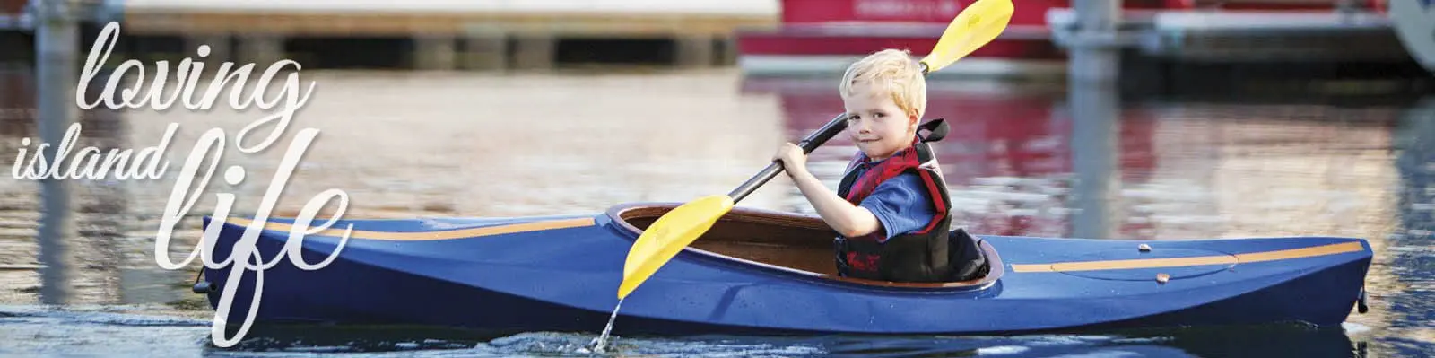 A child in a blue kayak holds a yellow paddle and navigates calm water. Text on the image reads "loving island life.