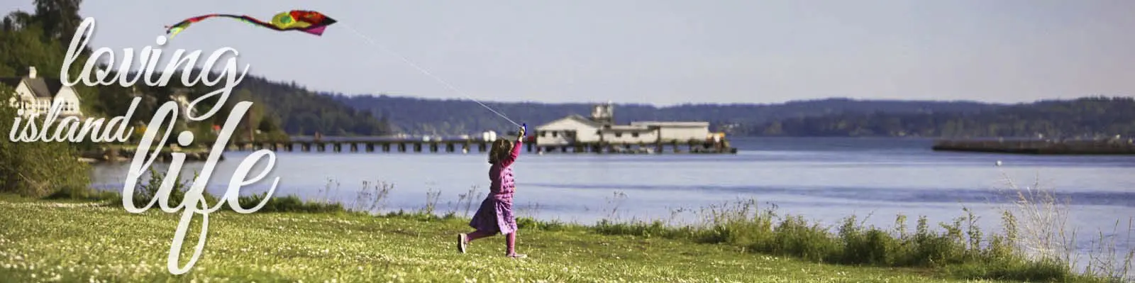 A child flies a kite on a grassy area by a waterfront, with a dock and buildings visible across the water. The text "loving island life" is displayed prominently on the left.