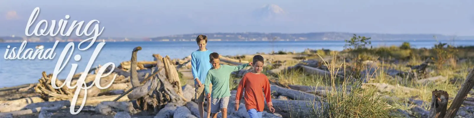 Three children walk along a beach with driftwood and sea grass, with mountains in the background. "Loving Island Life" is written on the left side of the image.