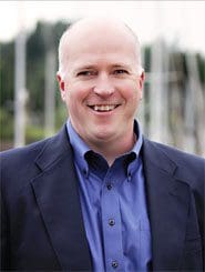 A smiling man in a blue shirt and dark blazer stands outdoors with blurred greenery in the background.