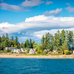 A waterfront view featuring a row of houses nestled among trees, with a backdrop of blue sky and scattered clouds.