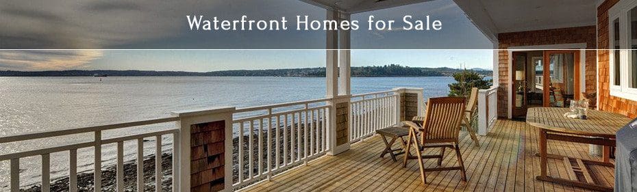 A spacious waterfront porch with wooden furniture overlooks a serene body of water under overcast skies. Text reads "Waterfront Homes for Sale.