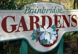 Sign reading "Bainbridge Gardens" with floral illustration, displayed outdoors against a backdrop of greenery.