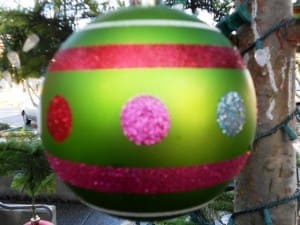 Close-up of a green Christmas ornament with horizontal red stripes and pink, red, and blue glitter dots. Trees and outdoor decorations are visible in the background.