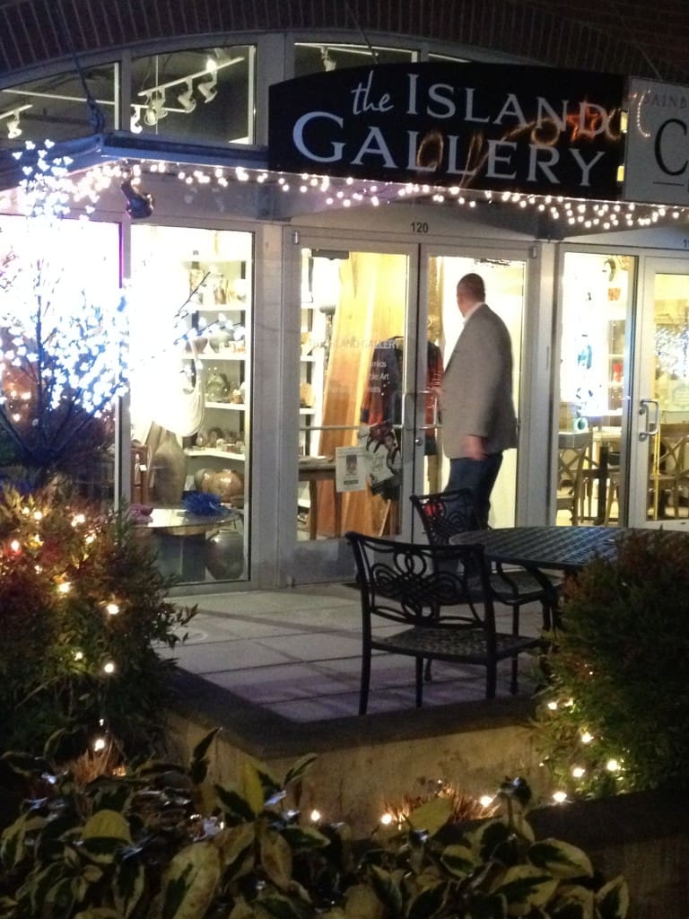 A man in a blazer stands at the entrance of The Island Gallery, a storefront decorated with lights and featuring a patio table and chairs and greenery in the foreground.