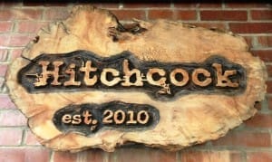 Wooden sign on a brick wall with the text "Hitchcock est. 2010" carved into it.