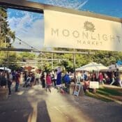 People are walking and shopping at an outdoor market called Moonlight Market, with various stalls and tents set up under a clear sky.