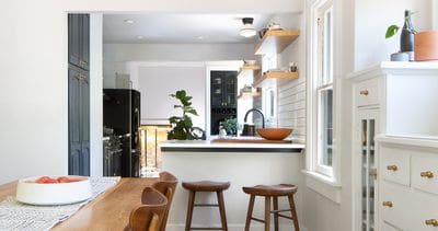 Bright kitchen with a wooden table, three wooden stools at the counter, and floating shelves over a tile backsplash. A white cabinet and a black refrigerator are visible in the background.