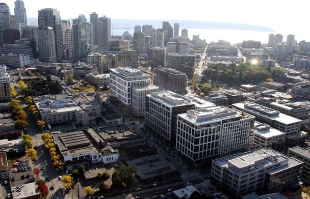 Aerial view of a cityscape with modern high-rise buildings, streets lined with trees showing fall colors, and a waterfront in the background under a bright sky.