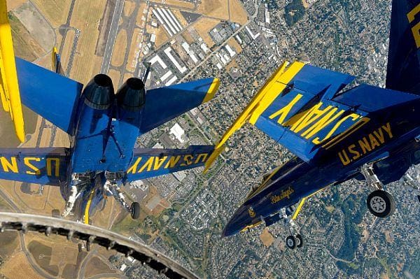 Two U.S. Navy Blue Angels jet aircraft fly upside down in close formation above a city landscape near an airfield.