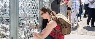 A person wearing a red top and backpack crouches to attach a lock to a fence covered with locks on a bridge, while other people walk by in the background.