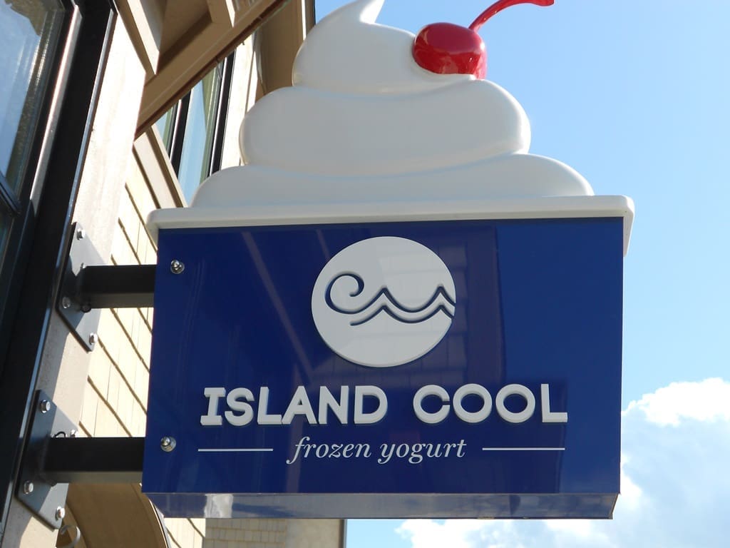 Blue and white "Island Cool frozen yogurt" sign with a swirl of frozen yogurt and a cherry on top, mounted on a building wall.