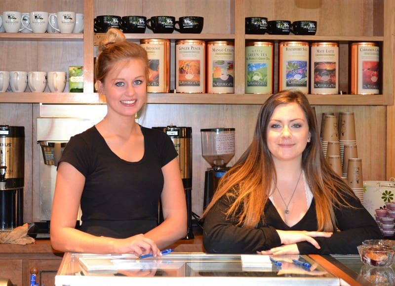 Two women standing behind a counter in a coffee shop, with various tea containers on a shelf behind them. One has light hair pulled back and is wearing a black shirt, the other has long dark hair and a black top.