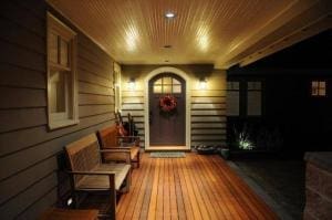 A well-lit wooden porch at night with a bench, guitar, and a front door decorated with a wreath.