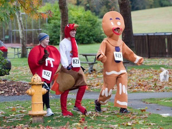 Three people are participating in a fun run wearing costumes: a heart, a turkey, and a gingerbread man. They have race bibs on with numbers 822, 821, and 823 respectively.