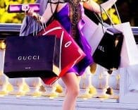 A person in a purple outfit carrying multiple branded shopping bags, including Gucci and Chanel, while walking outdoors.