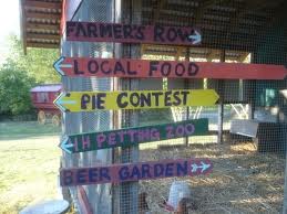 Colorful wooden signs on a farm point to various attractions: Farmers Row, Local Food, Pie Contest, 1H Petting Zoo, and Beer Garden.