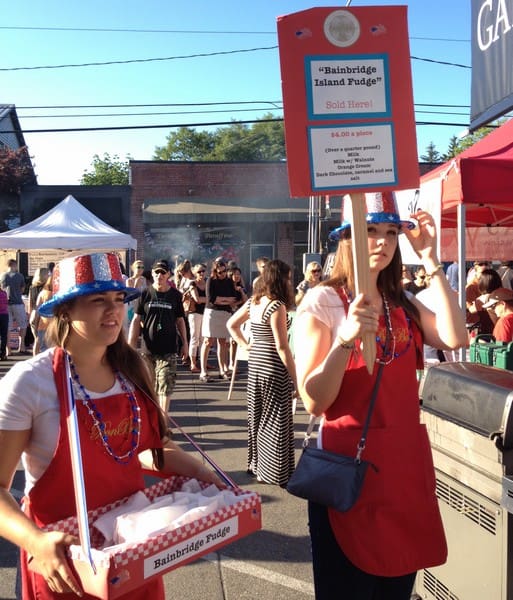 Two women dressed in festive attire sell fudge at an outdoor event. One holds a tray of fudge, while the other carries a sign advertising "Bainbridge Island Fudge" for $4.00 a piece.