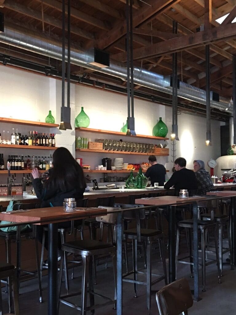 Interior of a bar with a wooden ceiling and industrial lighting. The bar is stocked with bottles and glasses, while several patrons sit on barstools.