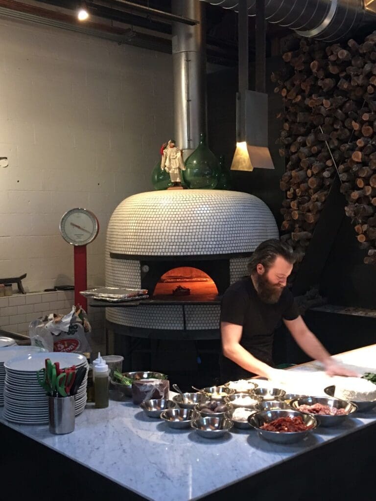 A man is preparing food on a marble countertop in a kitchen with various ingredients in metal bowls. Behind him is a large, tiled pizza oven with visible fire. A stack of firewood is in the background.