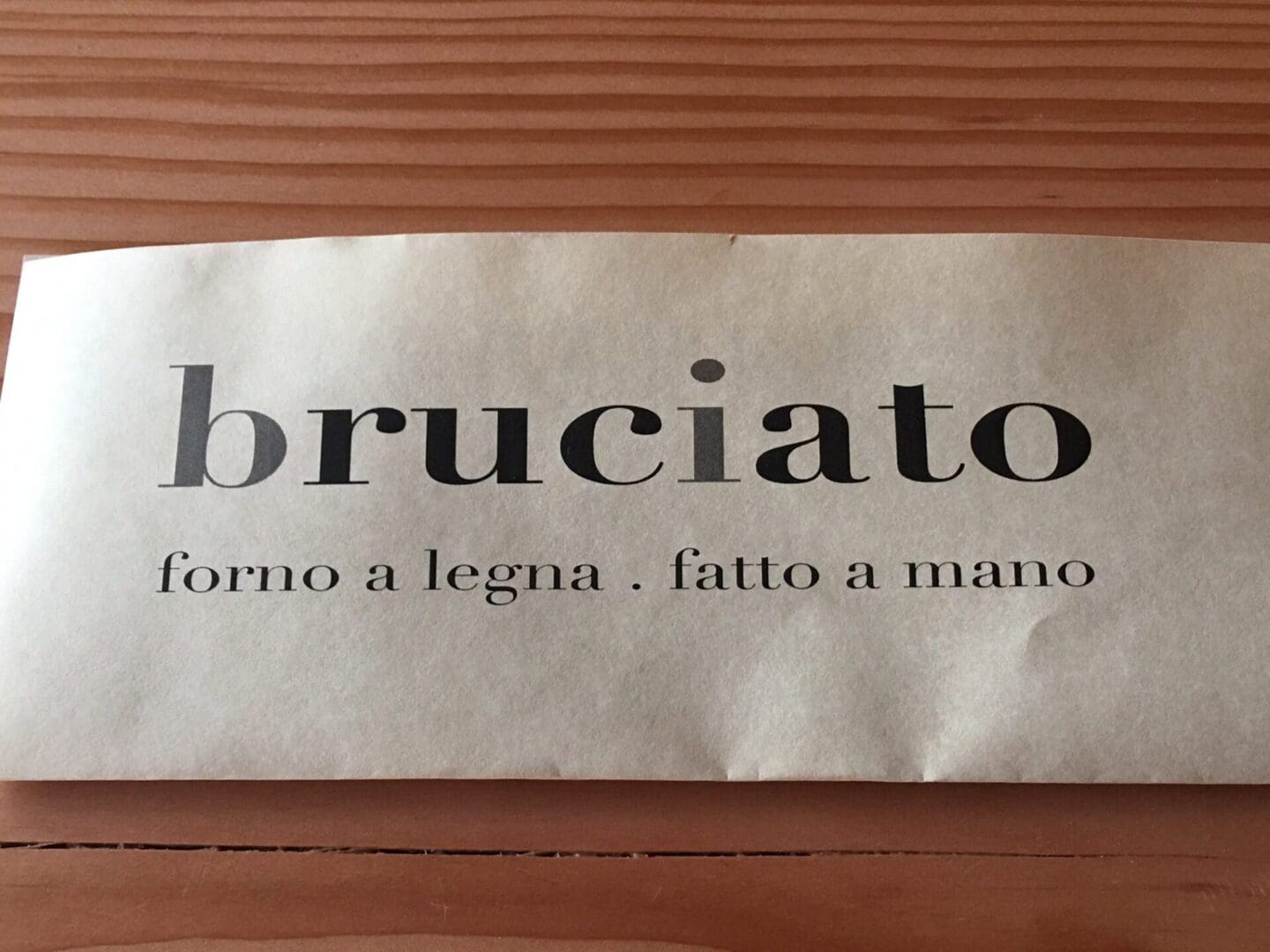 A paper with the text "bruciato forno a legna . fatto a mano" placed on a wooden surface.