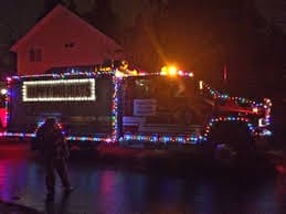 A fire truck decorated with colorful Christmas lights is parked on a wet street at night, with a dimly lit house in the background.