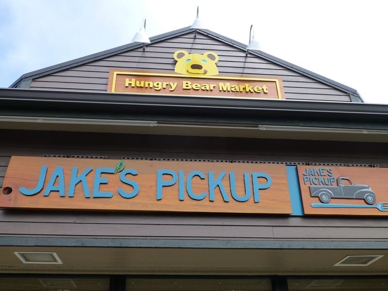 Image of the storefront signs for "Hungry Bear Market" and "Jake's Pickup" on a building. The Hungry Bear Market sign features a bear's face, and the Jake's Pickup sign includes an illustration of a truck.