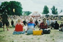 Three people sit on the grass, facing an outdoor stage with a crowd gathered in front of it. The scene is set in a park with trees and tents in the background.