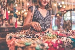 A woman with a light brown shoulder bag browses a table of colorful jewelry and accessories in a brightly lit market.