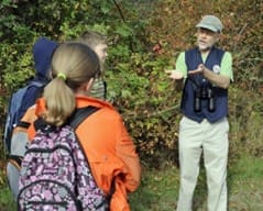 An older man wearing a cap talks to three children outdoors. One child wears a backpack with a floral pattern. The group is surrounded by greenery.