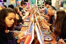 A group of people sit at tables, focused on painting canvases. Various colors and painting tools are visible.