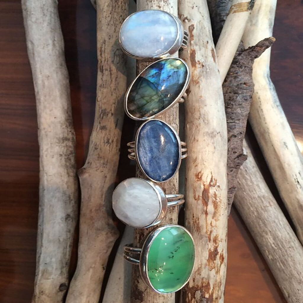 Five silver rings with large oval stones in different colors are displayed on wooden sticks: white, green, blue, brown, and a white stone with darker inclusions.