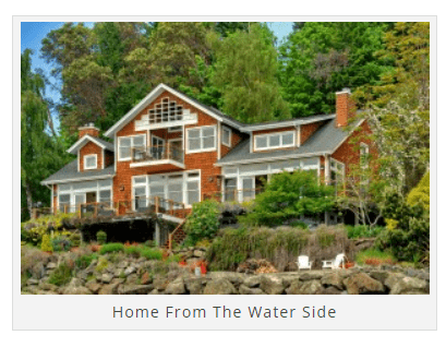 A large house with multiple stories, wooden exterior, and expansive windows. It is surrounded by trees and situated near a rocky waterfront. Two chairs and a small table are visible near the water.