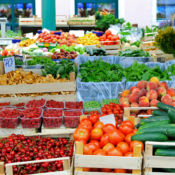 A colorful display of fresh fruits and vegetables at a market, including tomatoes, cucumbers, peaches, strawberries, and leafy greens, all arranged in wooden crates and baskets.