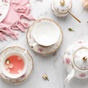 A floral teapot, a cup with pink tea, another empty cup, a sugar bowl, and a pink fringed cloth are arranged on a white marble surface with scattered flower petals.