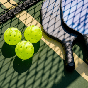 Two pickleball paddles and three yellow pickleballs are placed on a pickleball court, partially shadowed by the net.