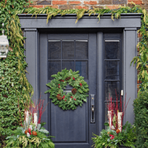 A dark blue front door decorated with a green wreath featuring pinecones, flanked by two planters with red twigs, white birch branches, and greenery. The doorway is framed with hanging greenery.