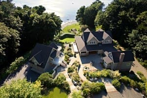 Aerial view of two houses with dark roofs surrounded by trees, near a body of water. A driveway and landscaped garden are visible between the houses.