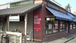 Street view of a brick building with two shops. The left shop is "Eagle Harbor Book Co." with a "Books" sign. The right shop has a large red sign that reads, "EAT SLEEP READ." Blue awning above windows.