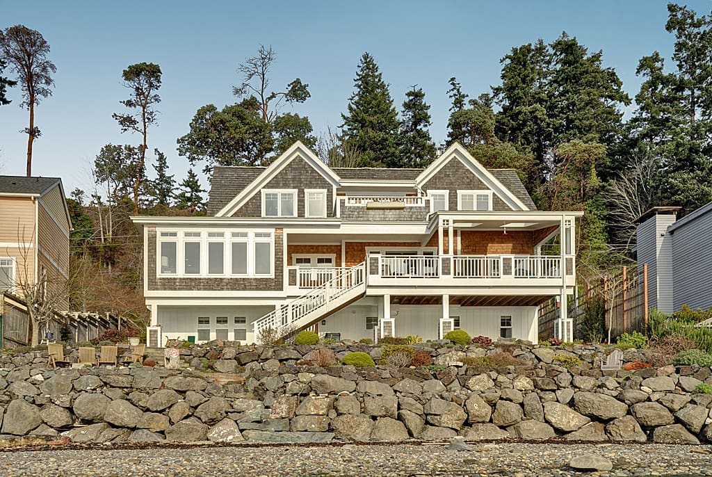 A two-story house with large windows and a spacious deck, surrounded by trees and built on a stone retaining wall.